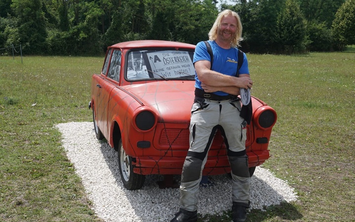 Ein Mann mit langen blonden Haaren, einer Motorradhose und einem blauen T-Shirt steht an eine rote Trabbi-Statue gelehnt