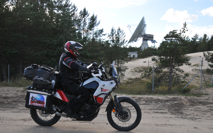 rot-weisses Motorrad auf einem Sandweg; im Hintergrund ist eine grosse Satellitenschuessel zu erkennen