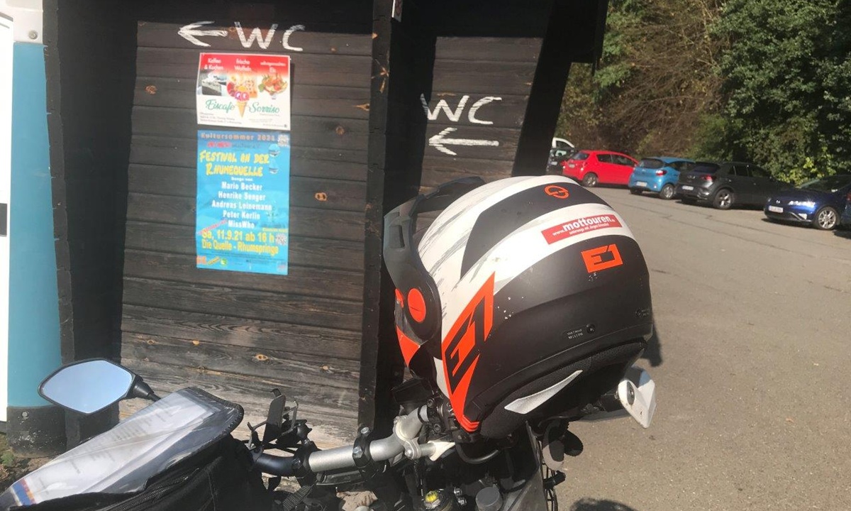 auf dem Lenker eines Motorrads liegender Motorradhelm; im Hintergrund ein auf ein WC hinweisendes Schild