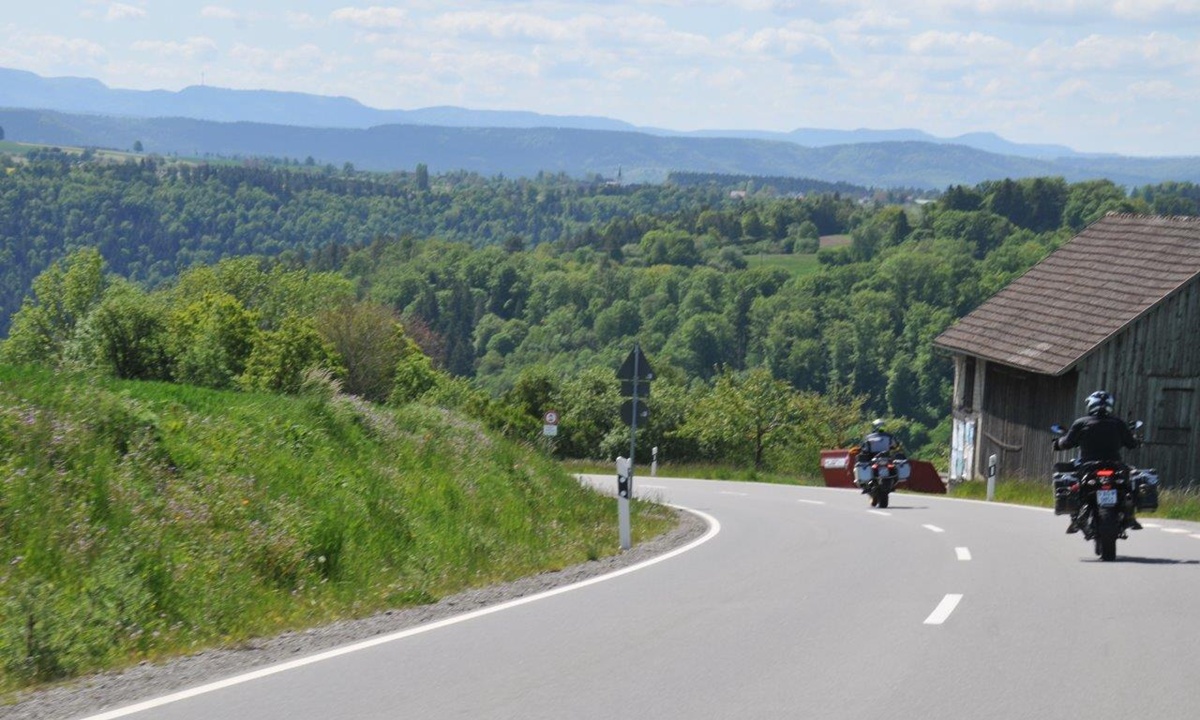 Motorraeder fahren auf einen Wald zu; am rechten Strassenrand steht ein Holzschuppen