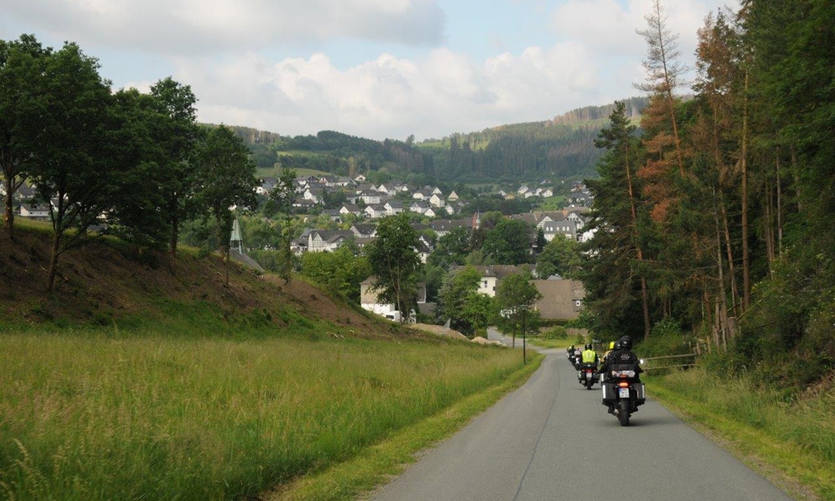 Drei Motorraeder fahren auf einer in eine Siedlung fuehrende Strasse
