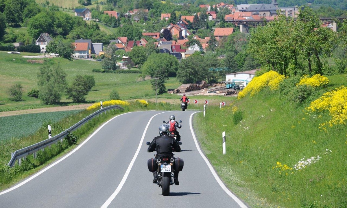 mehrere Motorraeder fahren auf einer Landstrasse zwischen Wiesen entlang auf eine Ortschaft zu