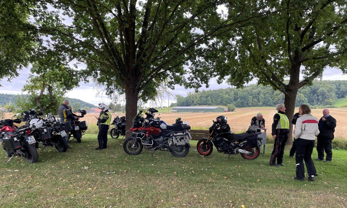 Eine Gruppe Motorradfahrer macht Pause im Schatten einiger grosser Baeume