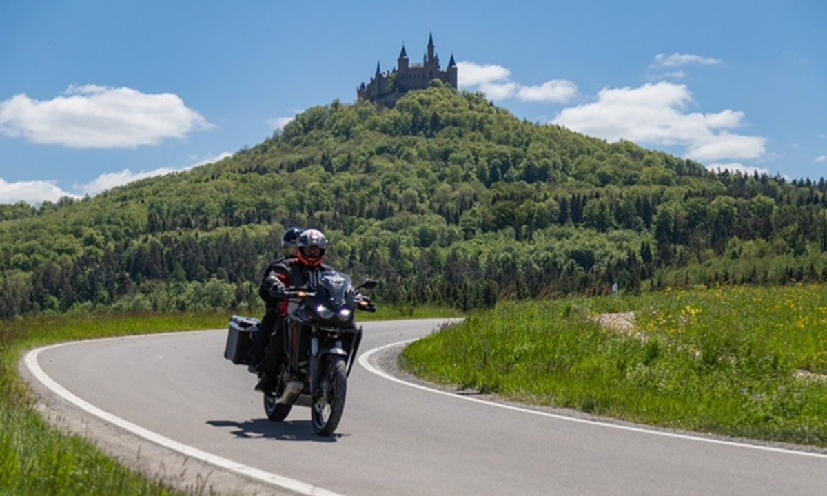 ein Motorrad fahrt eine Strasse entlang; im Hintergrund befindet sich ein Berg auf dessen Spitze eine Burg trohnt