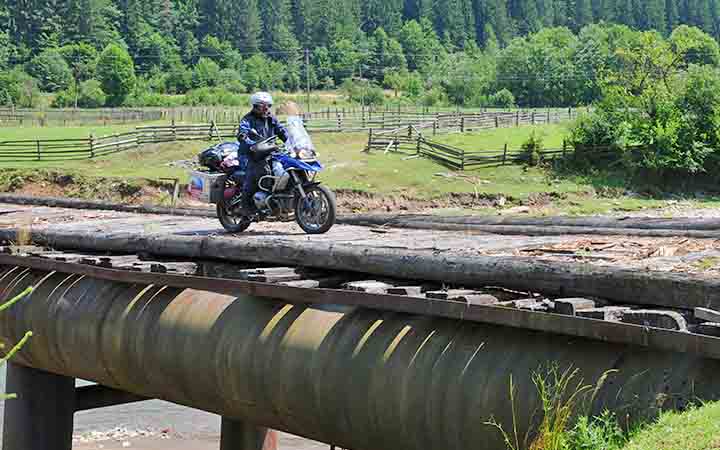 ein Motorrad faehrt auf einer alten im industriellen Stil erbauten Bruecke ueber einen Fluss in einer bergigen Waldlandschaft