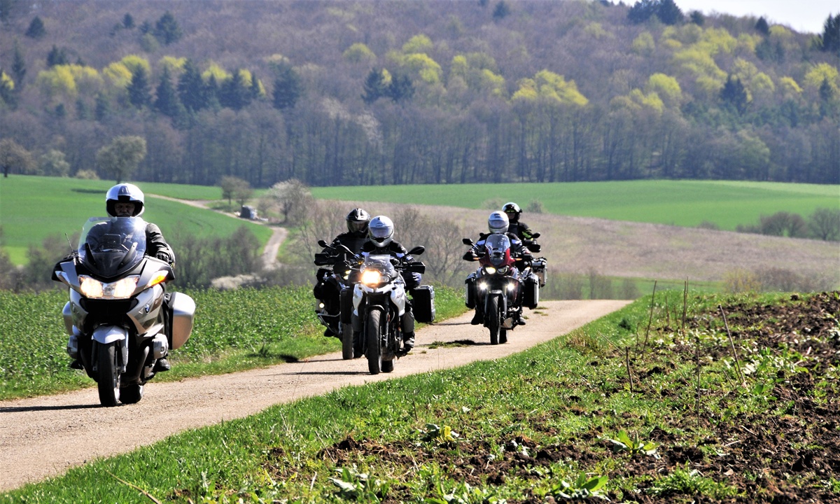 eine Gruppe Motorradfahrer faehrt ueber einen Feldweg; im Hintergrund befindet sich eine Waldlandschaft