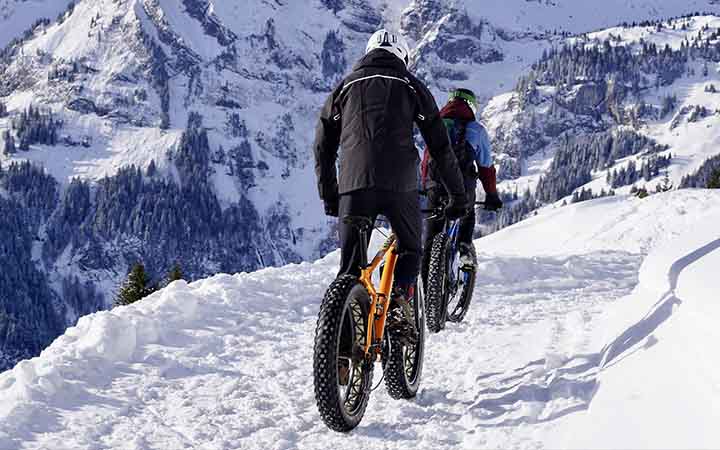 zwei junge Menschen fahren in Winterkleidung auf "Fatbikes" durch den Schnee; im Hintergrund sieht man einen verschneiten alpinen Berg
