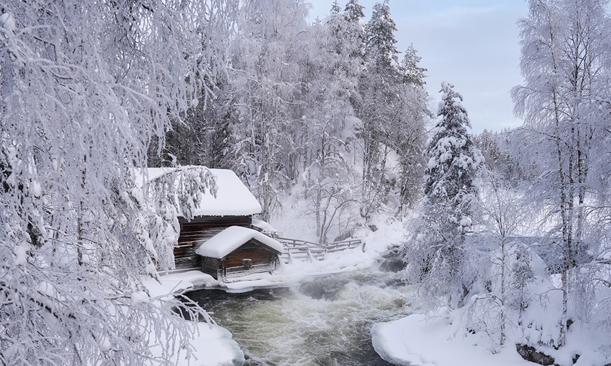 Eine kleine Holzhuette steht an einem Fluss in der verschneiten Winterlandschaft Lapplands